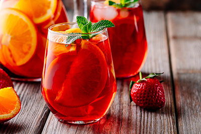 selbstgemixte Drinks mit Orangenscheiben und frischer Minze garniert. Mehrere bauchige Saftgläser stehen auf einem Gartentisch aus Holz. Eine Erdbeere liegt neben einem der Gäser.